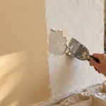 Рука со шпателем снимает старое покрытие с участка стены рядом с бежевой окрашенной поверхностью